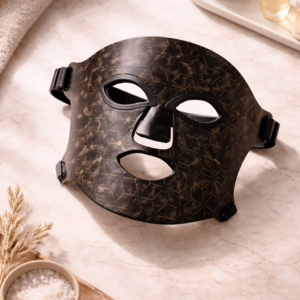 Dermaheal Pro LED Face Mask arranged in a flat-lay on a light marble surface, featuring a matte black finish with subtle gold geometric detailing, surrounded by soft spa elements like skincare jars, serum bottles, fabric, and natural accents in warm, calming tones.